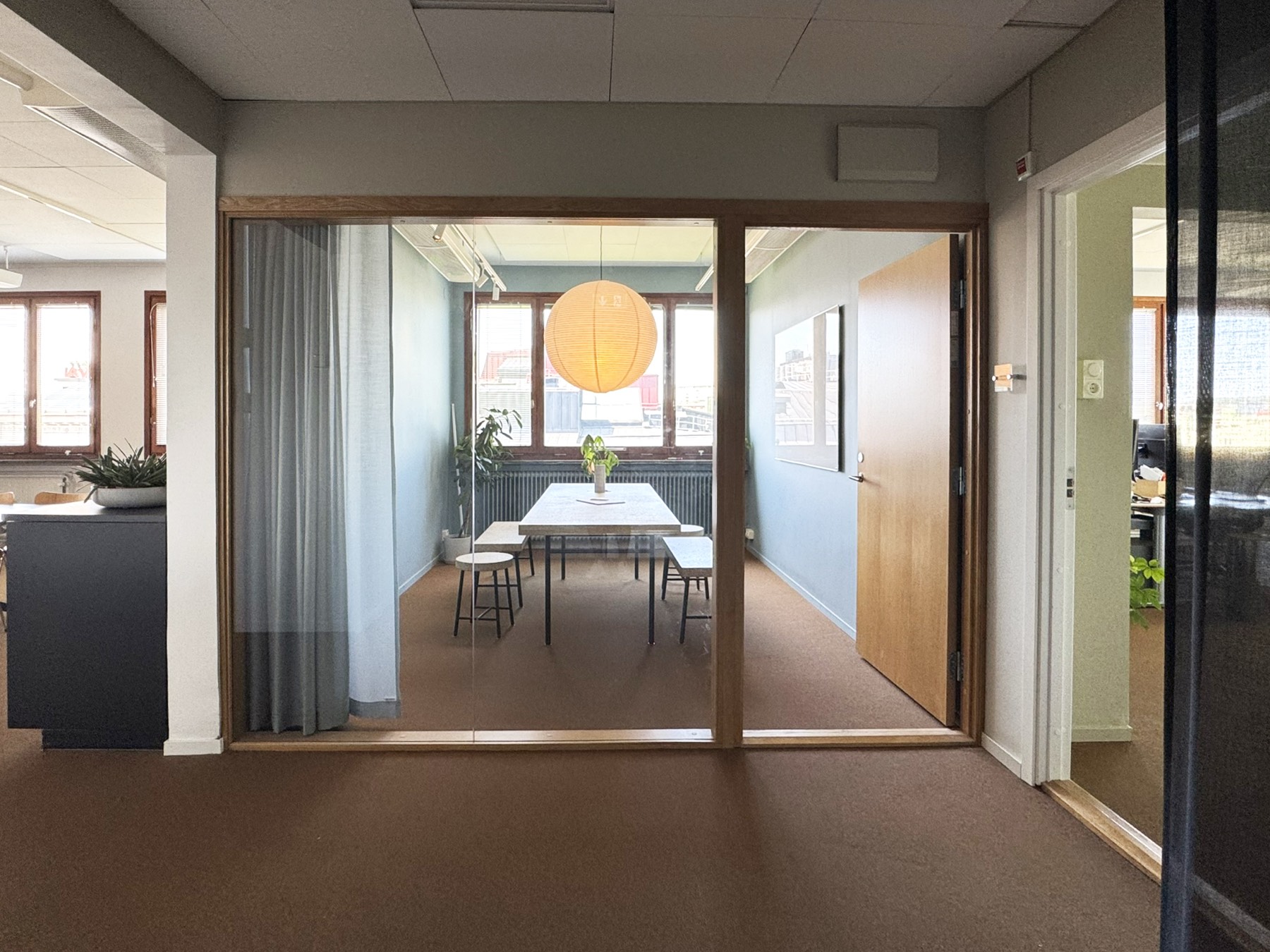 Entré med glasparti i ek in mot mindre mötesrum med papperslampa.